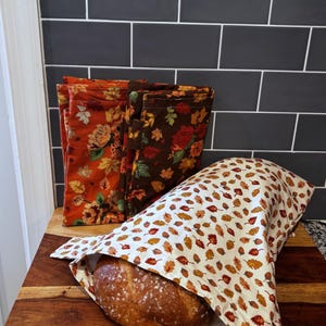 May include: A round loaf of crusty bread partially covered by a white fabric bag featuring a pattern of small autumn leaves and acorns, with two additional folded fabric bags in orange and brown floral leaf prints standing upright behind it.