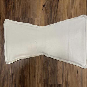 May include: A cream-colored, rectangular pillow with a unique bow-tie shape. The pillow has a stitched border and is set against a wooden background. The pillow's design is simple and minimalist.