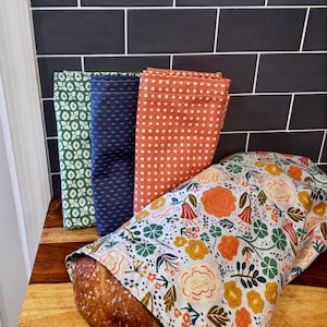 May include: A collection of four fabric wraps, including three folded rectangular cloths in green with white patterns, navy blue with white dashes, and orange with white checks, plus one floral print cloth covering a round loaf of bread.