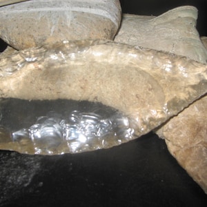 May include: A clear, translucent arrowhead-shaped piece of flint or obsidian, likely used as a tool or weapon in the past. It is resting on a piece of rough, brown leather or hide.