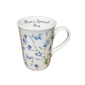 Puede incluir: Taza de cerámica blanca con un diseño floral de campanillas azules y tallos verdes. La taza tiene un asa curvada y el texto "Mum's Special Mug" en el borde.