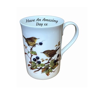 May include: White ceramic mug with a curved handle, decorated with an illustration of two small birds on a branch with berries and leaves. The mug has the text "Have An Amazing Day xx" inside the rim.