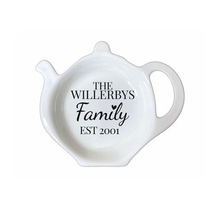 Op de afbeelding: Witte keramische theepotvormige lepelhouder met een handvat en tuit. De lepelhouder heeft de tekst "THE WILLERBYS Family EST 2001" in zwart schrift. Een decoratief en functioneel keukenaccessoire.