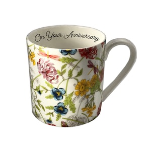 Puede incluir: Taza de cerámica blanca con un estampado floral con flores rojas, rosas, amarillas y azules, hojas verdes y libélulas. Las palabras "On Your Anniversary" están impresas dentro del borde de la taza.