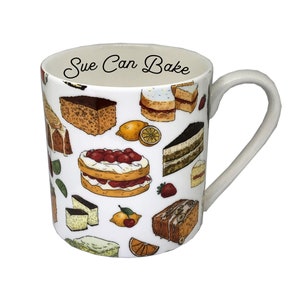 Puede incluir: Taza de cerámica blanca con asa, decorada con ilustraciones coloridas de varios pasteles, frutas y el texto "Sue Can Bake". La taza presenta una variedad de productos horneados, incluyendo pasteles en capas, rebanadas de pastel y fruta.
