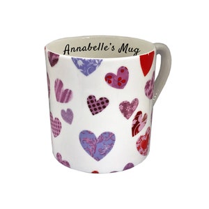 Może przedstawiać: Biały ceramiczny kubek ozdobiony różnymi wzorami serc w odcieniach czerwieni, fioletu i różu. Kubek ma szary uchwyt i napis "Annabelle's Mug" wewnątrz obręczy. Serca mają różne tekstury i wzory.