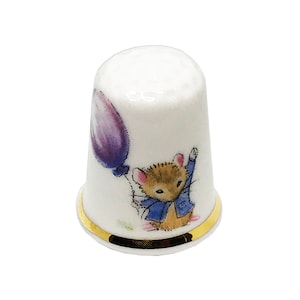 May include: White porcelain thimble with a gold band at the base. The thimble features a cartoon illustration of a mouse holding a purple balloon. The mouse is wearing a blue jacket. The thimble is a sewing accessory.
