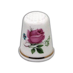 May include: White porcelain thimble with a pink rose design, green leaves, and blue accents. It has a gold band at the base and a dimpled top. A traditional sewing tool.