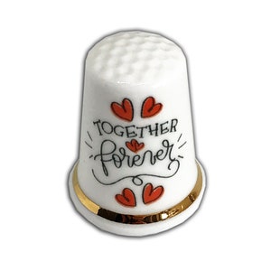 May include: A white ceramic thimble with a gold band at the base. The thimble is decorated with red hearts and the words "TOGETHER forever" in black script. The thimble has a dimpled texture.