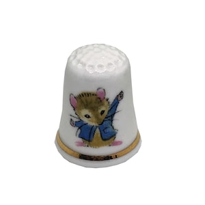 May include: White ceramic thimble with a gold band at the base. The thimble features a cartoon illustration of a mouse wearing a blue jacket. The top of the thimble has small, evenly spaced holes.
