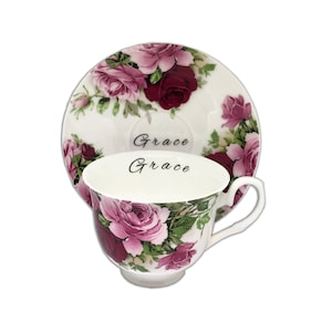 Op de afbeelding: Een witte theekop en schotel set met rozen in roze en bordeaux tinten. Het woord "Grace" staat in script op de kop en schotel. De theekop heeft een gebogen handvat. Een klassieke theeset.