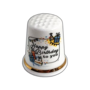 Puede incluir: Dedal de cerámica blanca con borde dorado, con el texto "Happy Birthday to you" con ilustraciones coloridas de regalos y decoraciones de fiesta. El dedal tiene una parte superior texturizada.