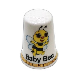 Puede incluir: Dedal de cerámica blanca con un diseño de abeja bebé de dibujos animados. La abeja tiene un cuerpo amarillo, rayas negras y alas azules. El dedal tiene una banda dorada en la base y las palabras "Baby Bee" están impresas debajo.