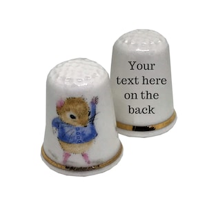 May include: Two white porcelain thimbles with gold trim. One thimble features a cartoon mouse wearing a blue jacket. The other thimble has the text "Your text here on the back".