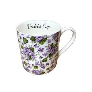 May include: White ceramic mug with a floral pattern of purple violets and green leaves. The mug has a curved handle and the text "Violet's Cup" in black script on the rim. The mug is a standard size.