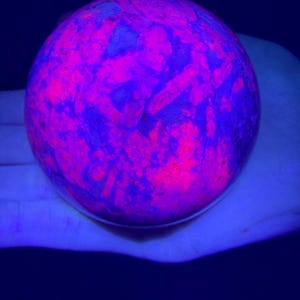 May include: A spherical stone displaying intense fluorescent properties under ultraviolet light, featuring a mottled pattern of vibrant neon pink and deep purple hues across its smooth, rounded surface, creating a high-contrast glowing effect.