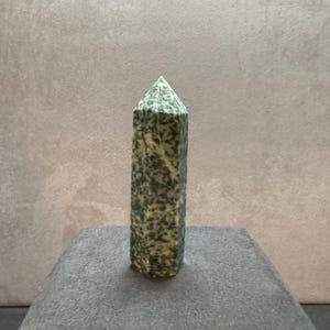 May include: A green and white speckled stone obelisk. The obelisk is rectangular with a pointed top. The stone has a polished finish and is displayed on a gray surface. The obelisk is a decorative item.