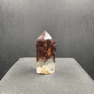 May include: A polished crystal point featuring a clear base that transitions into a deep, dark reddish-brown inclusion. The hexagonal tower has a pointed top and displays natural mineral patterns within the translucent and opaque sections.