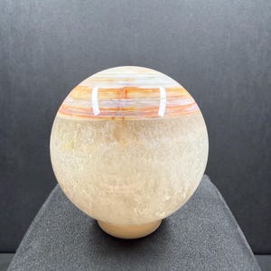 May include: A polished spherical stone object featuring a translucent, textured lower section in pale cream and a smooth upper band with horizontal layers of orange, white, and brown. The sphere rests on a small, circular base.