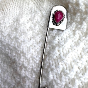 May include: A silver safety pin with a decorative head featuring a heart-shaped, faceted, red gemstone. The pin is set against a textured, white knitted background, highlighting the jewelry's design and color.