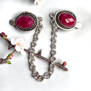 May include: Silver-toned shirt clips linked by a chain. Each clip has an oval, faceted, ruby-red gemstone in a decorative border. The clips are on a white surface with a sprig of white flowers.