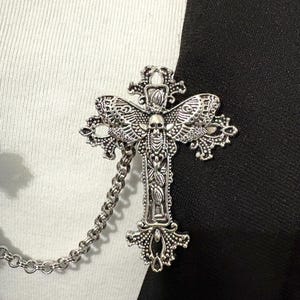Peut inclure: Broche en forme de croix argent&eacute;e avec des d&eacute;tails complexes, notamment un cr&acirc;ne et des ailes. La broche est reli&eacute;e &agrave; une cha&icirc;ne. Le fond est divis&eacute; en sections noires et blanches, cr&eacute;ant un contraste saisissant.