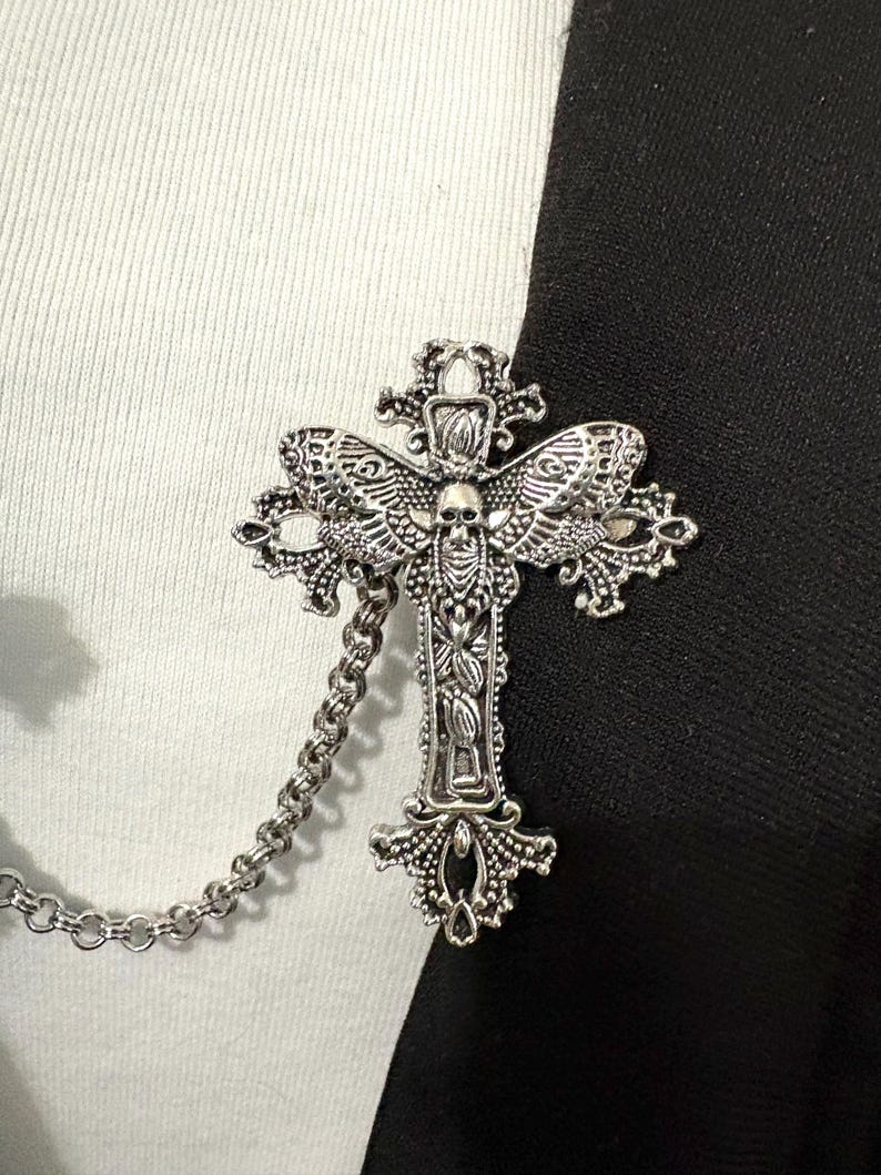 Peut inclure: Broche en forme de croix argent&eacute;e avec des d&eacute;tails complexes, notamment un cr&acirc;ne et des ailes. La broche est reli&eacute;e &agrave; une cha&icirc;ne. Le fond est divis&eacute; en sections noires et blanches, cr&eacute;ant un contraste saisissant.
