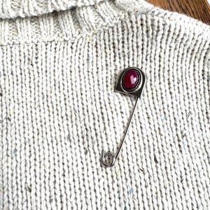 May include: A silver-toned brooch pin with a rectangular head featuring a faceted, oval-shaped red gemstone. The pin is attached to a light gray, textured knit sweater with a turtleneck collar.