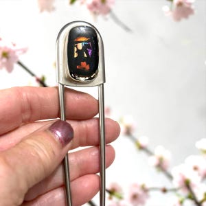 May include: A silver-toned metal hair fork featuring a rectangular black cabochon at the top. The cabochon displays iridescent, multi-colored flecks in shades of orange, purple, and blue that shimmer against the dark, glossy surface.