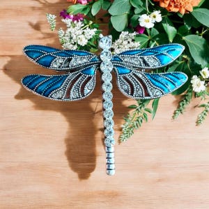 Peut inclure: Une broche en forme de libellule bleue et argentée avec des strass transparents. La libellule présente des détails complexes et un corps long et fin.