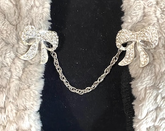 May include: Silver-toned bow-shaped brooch with clear crystal accents, connected by a delicate chain. The brooch is designed to fasten two sides of a garment together. The background is a black fabric and a fluffy, light gray material.