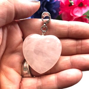 May include: A pale pink heart-shaped rose quartz pendant with a silver bail. The pendant is held in a hand.