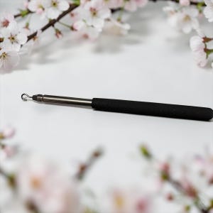 May include: A silver-coloured tool with a curved hook on one end and a black, cylindrical handle. The tool is designed for precision work. The background features white and pink cherry blossom branches.