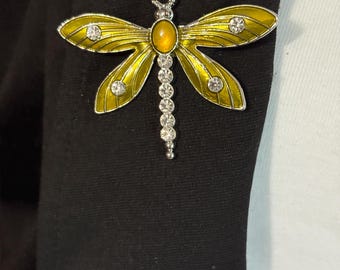 May include: A yellow and silver dragonfly brooch with clear rhinestone accents. The dragonfly has a yellow body and wings, with silver detailing. The brooch is attached to a black fabric.