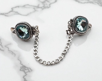 May include: A silver brooch featuring two square, pale blue gemstones linked by a chain. Each gem is set within a decorative, textured frame. The brooch is presented on a white and grey marble surface.