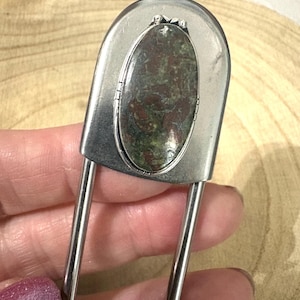 May include: A large silver-toned safety pin brooch featuring an oval-shaped stone with mottled green and deep red coloring set in a polished metal frame. The stone is secured with small metal prongs at the top and bottom of the setting.