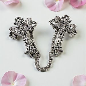 Peut inclure: Broche argent&eacute;e en forme de croix, reli&eacute;e par une cha&icirc;ne. Les croix pr&eacute;sentent des d&eacute;tails complexes et des &eacute;l&eacute;ments d&eacute;coratifs. Des p&eacute;tales de fleurs roses sont dispers&eacute;s autour de la broche sur fond blanc.