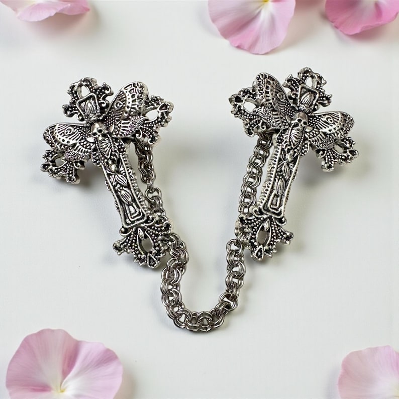 Peut inclure: Broche argent&eacute;e en forme de croix, reli&eacute;e par une cha&icirc;ne. Les croix pr&eacute;sentent des d&eacute;tails complexes et des &eacute;l&eacute;ments d&eacute;coratifs. Des p&eacute;tales de fleurs roses sont dispers&eacute;s autour de la broche sur fond blanc.