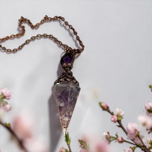 Peut inclure: Un collier pendentif composé d'un cristal d'améthyste en forme de cône pointu, enchâssé dans un capuchon en métal cuivré vieilli aux détails filigranés complexes, suspendu à une chaîne à maillons en métal cuivré assortie.