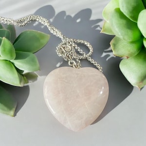 May include: A delicate silver chain necklace with a large, heart-shaped rose quartz pendant. The pendant is smooth and translucent, with a pale pink hue.