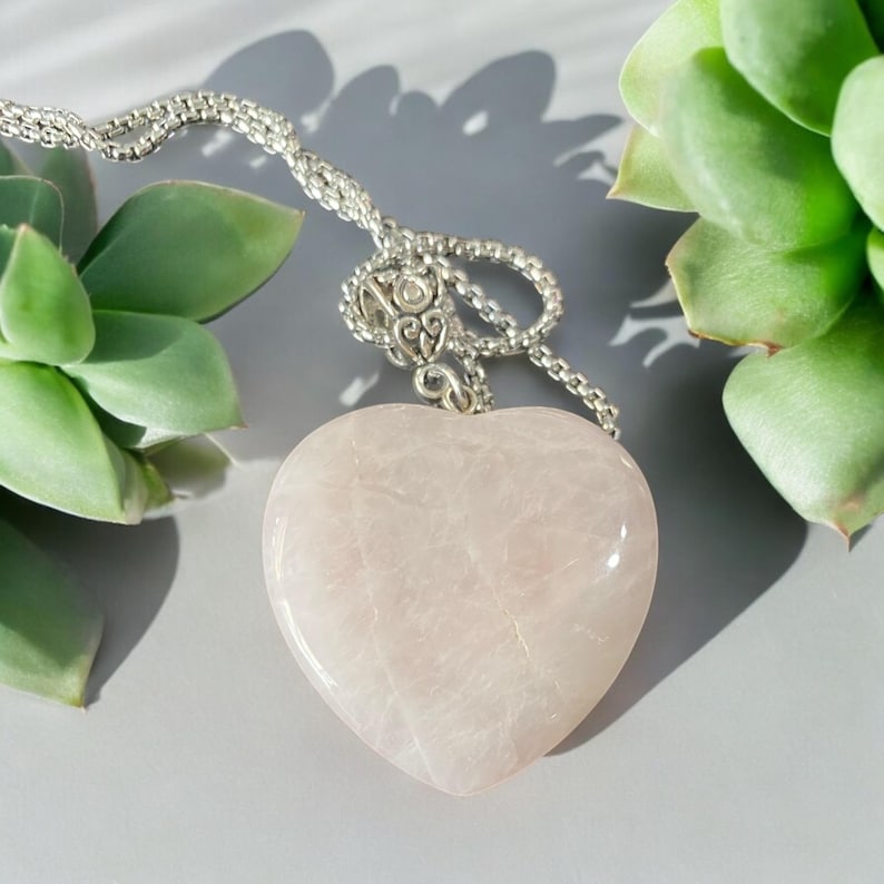 May include: A delicate silver chain necklace with a large, heart-shaped rose quartz pendant. The pendant is smooth and translucent, with a pale pink hue.