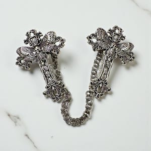 Peut inclure: Broche argent&eacute;e en forme de croix avec des d&eacute;tails complexes et une cha&icirc;ne de connexion. La broche pr&eacute;sente des motifs orn&eacute;s et une esth&eacute;tique gothique. Les deux croix sont reli&eacute;es par une cha&icirc;ne.