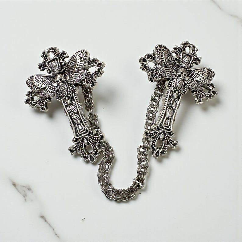 Peut inclure: Broche argent&eacute;e en forme de croix avec des d&eacute;tails complexes et une cha&icirc;ne de connexion. La broche pr&eacute;sente des motifs orn&eacute;s et une esth&eacute;tique gothique. Les deux croix sont reli&eacute;es par une cha&icirc;ne.