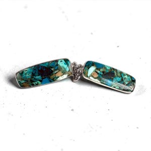 Peut inclure: Un bracelet argenté avec deux incrustations rectangulaires en pierre turquoise. Les pierres sont une mosaïque de teintes bleues, vertes et brunes. Les deux pièces rectangulaires sont reliées par une chaîne argentée.