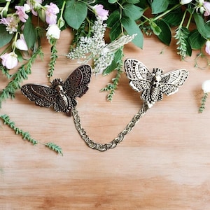 May include: Two silver butterfly brooches with a skull design connected by a silver chain. The brooches are detailed with intricate patterns and have a vintage aesthetic.