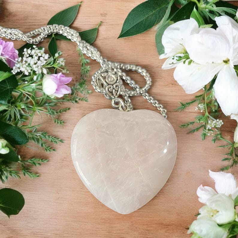 May include: A white heart-shaped pendant made of polished stone hangs from a silver chain. The pendant has a delicate, marbled texture. The chain is made of small, linked silver beads.