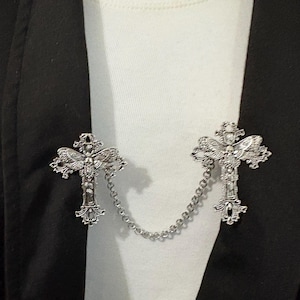 Peut inclure: Broche en forme de croix argent&eacute;e avec des d&eacute;tails orn&eacute;s, reli&eacute;e par une fine cha&icirc;ne. La broche est pr&eacute;sent&eacute;e sur une chemise blanche et un blazer noir, mettant en valeur son design et son utilisation comme accessoire de mode.