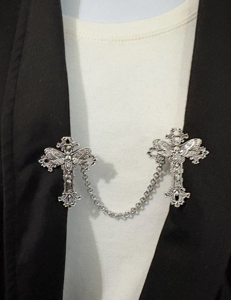 Peut inclure: Broche en forme de croix argent&eacute;e avec des d&eacute;tails orn&eacute;s, reli&eacute;e par une fine cha&icirc;ne. La broche est pr&eacute;sent&eacute;e sur une chemise blanche et un blazer noir, mettant en valeur son design et son utilisation comme accessoire de mode.