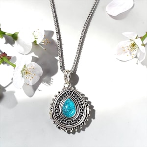May include: A silver necklace with a turquoise teardrop pendant. The pendant has an ornate, detailed design. White flowers are arranged around the necklace against a white backdrop.