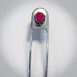 May include: A silver-toned hairpin with a teardrop-shaped, deep pink gemstone set in an ornate frame. The pin has a classic safety pin design, with a long, straight pin and a curved top.